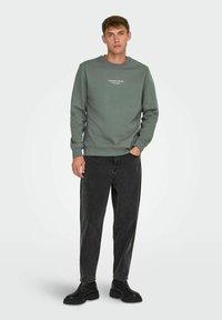 Heather grøn sweatshirt med rund hals og ribbede manchetter, parret med sorte løse jeans og sorte ankelstøvler. Simpelt design og tekstur.