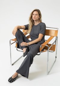 Grijze, gestructureerde jumpsuit met korte mouwen en uitlopende broekspijpen, gecombineerd met zwarte schoenen met zilveren studs. Zit op een stoel met metalen frame.