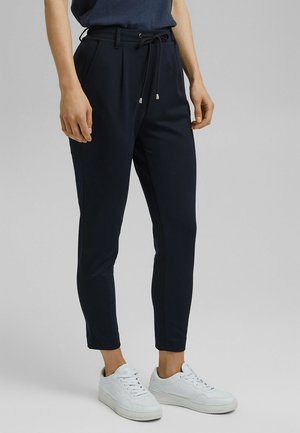 Osoba ubrana w granatowe, dopasowane spodnie do kostek z elastycznym pasem i sznurkiem oraz białe trampki, stojąca na tle jednolitego szarego tła.