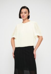 Femme aux cheveux courts et foncés portant un chemisier blanc à manches courtes avec bordure en dentelle et une jupe plissée noire taille haute sur fond blanc.