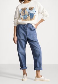 Žena v bílém svetru se dvěma plyšovými medvídky v džínách, modrých ohnutých kalhotách a bílých teniskách, stojící s jednou rukou v kapse.