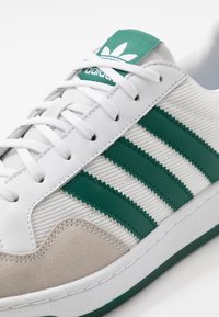 White sneaker featuring green accents, three green stripes, and a textured canvas upper with a beige suede toe cap.