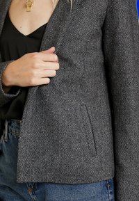Grauer Fischgrät-Blazer mit Reverskragen, einer einzelnen Tasche und tailliertem Design; getragen über einem schwarzen Oberteil und kombiniert mit Jeans.