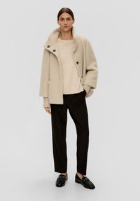 Beiger oversized Mantel mit großem Kragen und Knopfverschluss, über einem hellbeigen Pullover und schwarzen Hosen getragen, kombiniert mit schwarzen Mokassins.