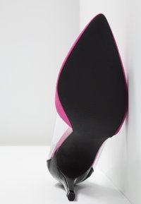 Chaussure à talon haut à bout pointu avec semelle noire, bord supérieur rose et côtés transparents, posée à la verticale contre une surface blanche.