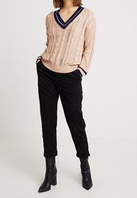 Donna che indossa un maglione beige a maglia con scollo a V e rifiniture blu navy, pantaloni neri arrotolati e stivaletti neri con tacco, in piedi con le mani in tasca.