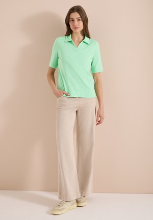 Frau steht mit einer Hand in der Tasche und trägt ein kurzärmeliges mintgrünes Polohemd, beige weite Hosen und beige Turnschuhe.