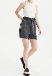 Pantaloni corti grigi con coulisse e bottoni a pressione laterali, con dettagli gialli, abbinati a un top corto bianco e sandali neri senza chiusura. Tessuto liscio.