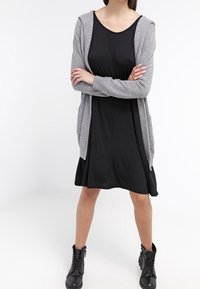 Cárdigan gris sobre un vestido negro, con un corte holgado y capucha en el hombro. Botines negros completan el look. Texturas suaves, diseño de longitud media.