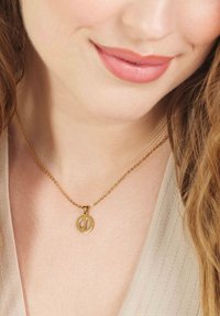 Gouden hangerketting met een circulair ontwerp en letterdetails op een delicate ketting, gedragen tegen een lichtgekleurde top.
