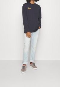 Person som bär en marinblå långärmad tröja med texten "LIFE AFFIRMING ACTIVISM", ljusblå uppvikta jeans, oranga strumpor och grå sneakers.