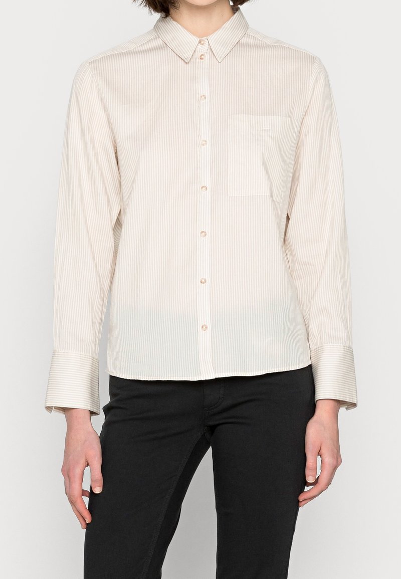 Personne portant une chemise beige à rayures avec une poche poitrine et un pantalon noir, debout devant un fond uni.