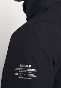 Detail černého rukávu bundy s logem značky ECOALF a textem o používání recyklovaných látek z kávových šálků pro ekologické produkty.