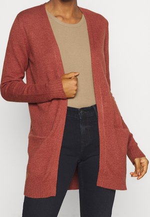 Personne portant un cardigan ouvert couleur rouille sur une chemise beige à manches longues et un pantalon noir taille haute, la main ajuste le cardigan.