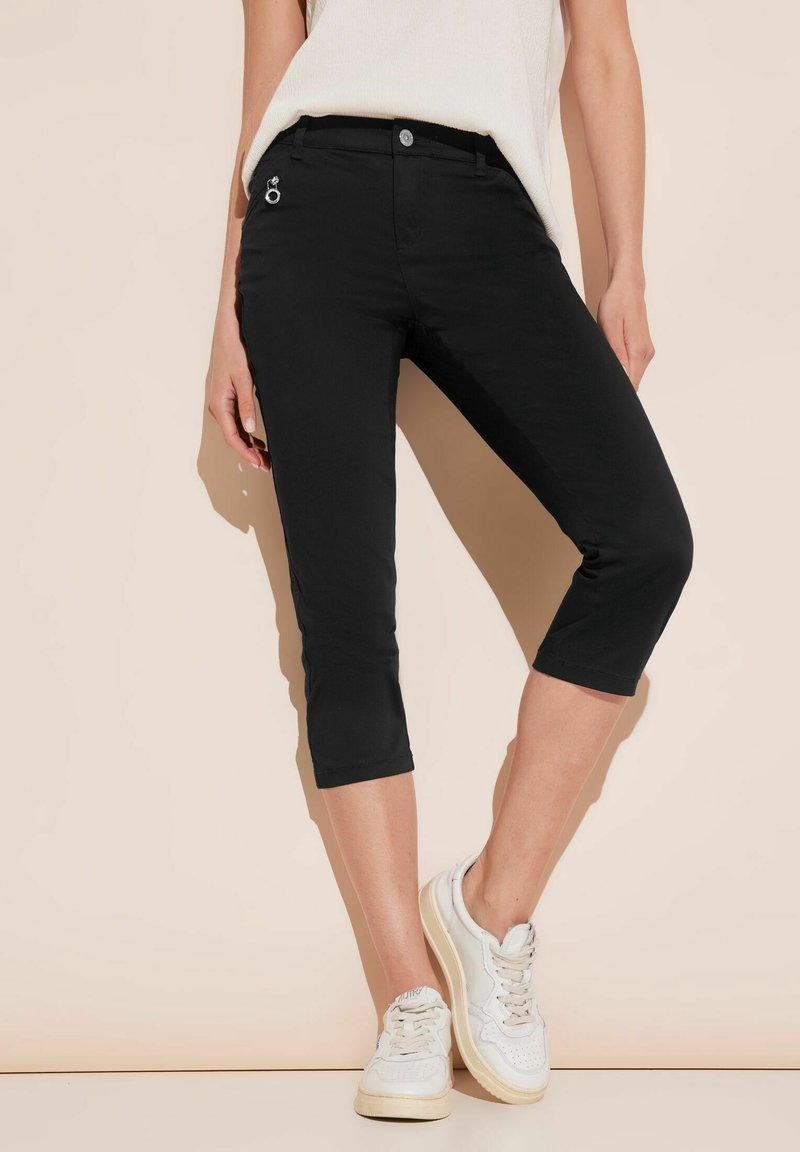 Femme portant un pantalon noir court avec une poche zippée à l'avant et des baskets blanches, debout contre un mur beige.