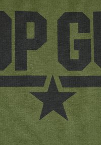 Olive green fabric featuring large black block letters spelling "TOP GUN" with a star and horizontal lines, textured knit material.