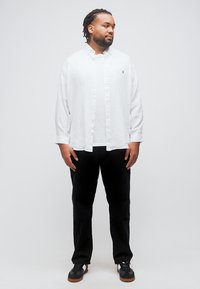 Άνδρας με πλεγμένα μαλλιά, φορώντας λευκό πουκάμισο με κουμπιά, μαύρο παντελόνι και μαύρα αθλητικά παπούτσια, στέκεται και κοιτάζει προς τα δεξιά σε απλό φόντο.