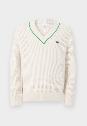 Pull en laine crème avec un col en V, des bordures vertes et des poignets côtelés. Présente un petit logo Lacoste sur la poitrine.