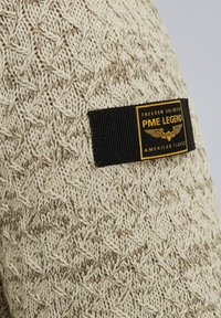Pull en tricot beige et crème avec un motif texturé. Présente une étiquette noire avec un texte jaune indiquant "PME LEGEND AMERICAN CLASSIC."