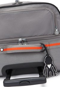 Valise à roulettes grise avec un tissu texturé, bande orange contrastante, doubles zips et une étiquette porte-clés en forme de singe noir attachée.