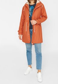 Parka orange résistant à l'eau avec une capuche, arborant des cordons blancs, une doublure crème douce et une coupe décontractée sur un jean bleu et des baskets blanches.