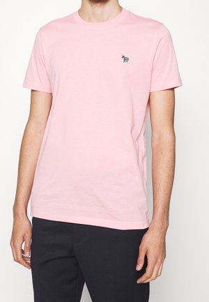 Mand iført simpel lyserød kortærmet t-shirt med lille zebra-logo på venstre bryst og sorte bukser mod hvid baggrund.
