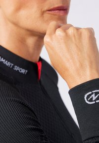 Schwarzes Sportoberteil mit roten Akzenten am Hals, aus strukturiertem Stoff und weißen Logodetails am Bündchen. Nahaufnahme einer Hand, die eine Faust bildet.