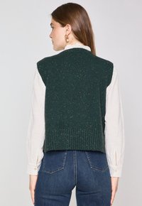 Gilet verde lavorato a maglia con superficie testurizzata e orlo a coste, indossato sopra una camicetta bianca, caratterizzato da ampie aperture per le braccia e un design corto.