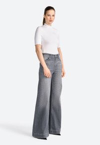 Hvid tætsiddende top med bølget halsudskæring, parret med højvirkende, brede grå jeans og sorte spidse høje hæle. Glat stof og rent design.