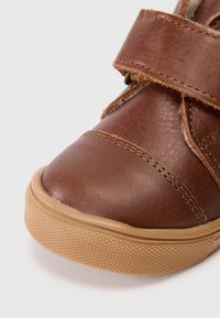 Sapato de couro castanho com bico arredondado, detalhes cosidos e sola de borracha texturizada. Apresenta uma fita de velcro para um fecho seguro.