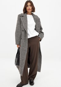 Lang, dobbeltradet frakke i houndstooth-mønster, grå og hvid, med store revers, båret over en hvid skjorte og brune bukser.