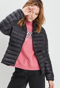 Zwarte pufferjack met horizontaal gestikt ontwerp en hoge kraag, gedragen over een roze sweatshirt. Eenvoudige ritssluiting en elastische manchetten.