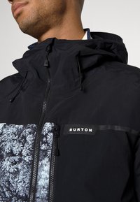 Veste noire avec une fermeture éclair à l'avant, une capuche et un panneau graphique avec des motifs d'arbres blancs. Étiquette logo sur la poitrine.