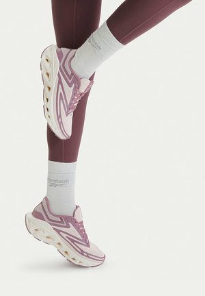 Piernas con leggings morados, calcetines blancos Reebok y zapatillas deportivas moradas y blancas con suelas gruesas levantadas contra un fondo claro.