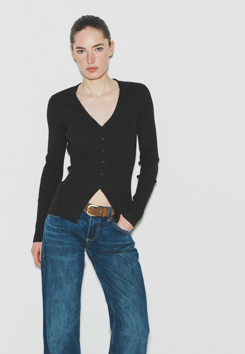 Vrouw met haar in een staart draagt een zwart V-hals top met knopen, blauwe jeans met een bruine riem, staand tegen een witte achtergrond met haar hand in haar zak.