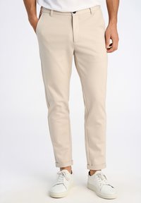 Pantaloni beige su misura con frontale piatto, tasche angolate e orli arrotolati, abbinati a sneakers bianche su uno sfondo neutro.