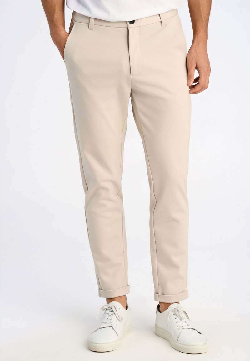 Pantaloni beige su misura con frontale piatto, tasche angolate e orli arrotolati, abbinati a sneakers bianche su uno sfondo neutro.