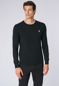 Maglione lungo nero a maniche lunghe con scollatura rotonda, bordi bianchi e un piccolo logo ricamato sul petto. Tessuto morbido e design aderente.