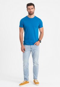 Modré tričko s kulatým výstřihem a krátkým rukávem, doplněné světlými modrými džíny a oranžovými teniskami. Hodinky na zápěstí, stojící na bílém pozadí.