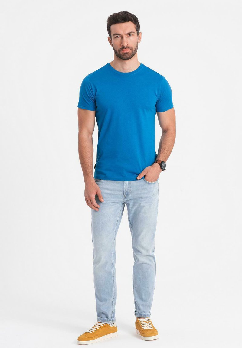 Modré tričko s kulatým výstřihem a krátkým rukávem, doplněné světlými modrými džíny a oranžovými teniskami. Hodinky na zápěstí, stojící na bílém pozadí.