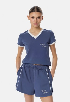 Naine kannab tumelsinist lühikesi pükse ja V-kaelusega valge äärega T-särki, millel on tekst „Cardio Bunny“, käed taskutes, neutraalne taust.