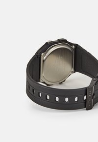 Montre numérique noire avec un cadran rond, bracelet en caoutchouc avec des trous, et un dos en acier inoxydable. Nom de la marque "Casio" sur le fermoir.