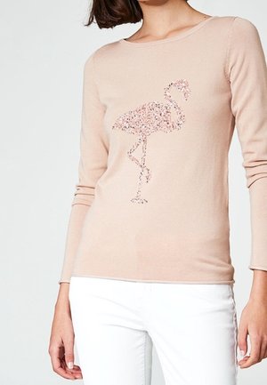 Vrouw in een lichtroze lange mouw trui met een pailletten flamingo ontwerp en witte broek, staande voor een effen achtergrond.