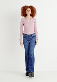 Cardigan rose clair à manches longues en tissu côtelé avec des détails de boutons, associé à un jean bleu évasé et des sabots noirs. Fond blanc uni.