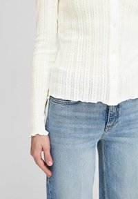 Biała, prążkowana bluzka z długim rękawem i zapięciem na guziki, z pionowymi paskami i małymi wzorami w kształcie oczek; zestawiona z jasnoniebieskimi dżinsami.