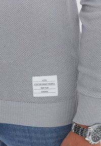 Helles graues Strickpullover mit gerippten Bündchen und Saum. Das Etikett auf der Unterseite lautet: "WÄHLT DIE RICHTIGEN PERSONEN, NICHT XXXXXXX."