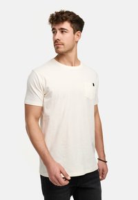 Cremefarbiges T-Shirt mit kurzen Ärmeln aus weichem Stoff, runder Ausschnitt und Brusttasche, mit einem kleinen Logo-Label an der Tasche.