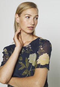 Blusa náutica floral com mangas curtas, com padrões de flores amarelas e verdes, textura suave e decote redondo.