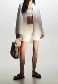 White button-up shirt, white cropped tank, and shorts. Multicolored crochet bag with a patterned design. Brown sandals with a buckle detail.