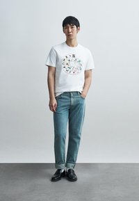 Λευκό βαμβακερό μπλουζάκι με πολύχρωμη γραφική εκτύπωση στο κέντρο. Φοριέται με ανοιχτό μπλε τζιν και μαύρα λόφερ.